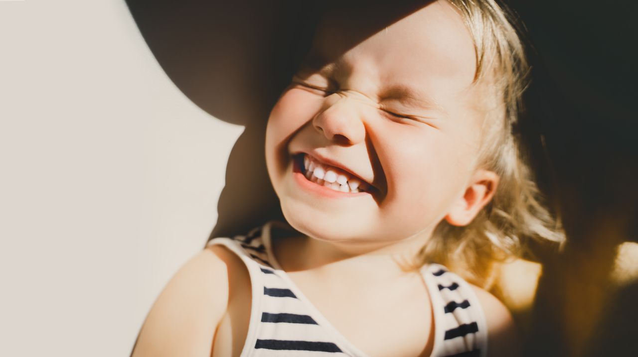 Photographie d'une petite fille qui sourit de manière exagérée en montrant toutes ses dents et en fermant les yeux.